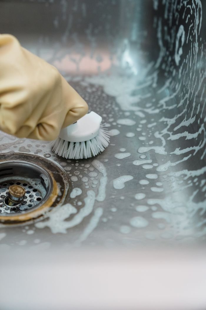 Home Close-up of a hand in gloves scrubbing a soapy kitchen sink with a brush.